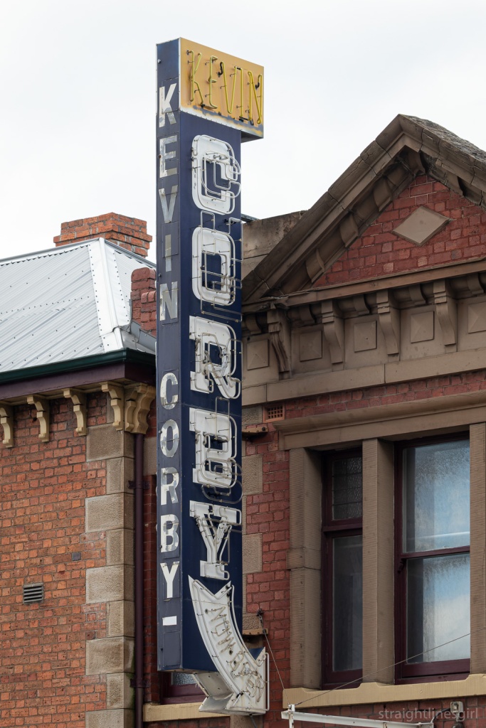 An old neon sign with the word KEVIN in yellow, horizontal above the word CORBY on blue vertical. It is in front of a brick building