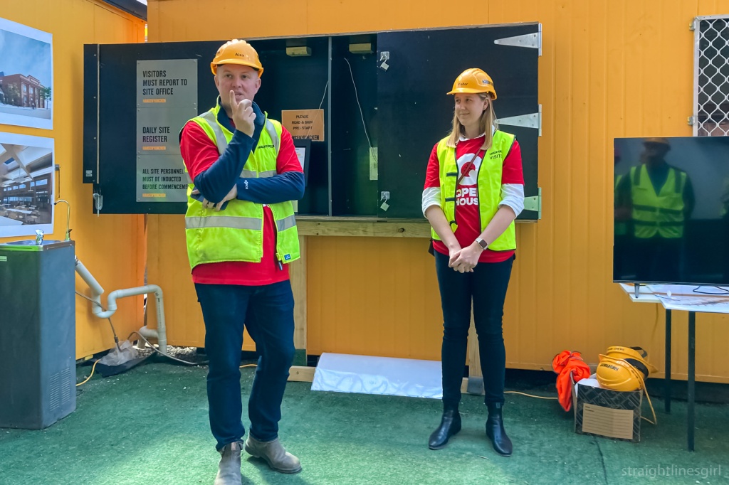 Two people dressed in black pants, red t0shirt, green hi vis vests and yellow construction hard hats standing in front of a yellow wall giving a talk
