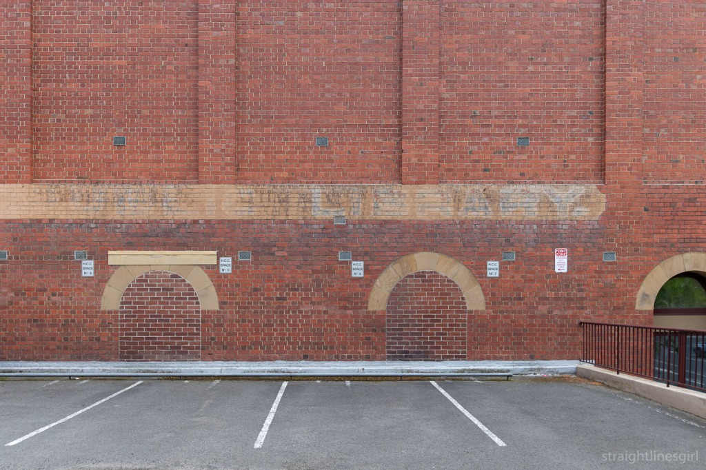 A red brick wall in a car park bearing a faded sign with the words PUBLIC LIBRARY faintly visible