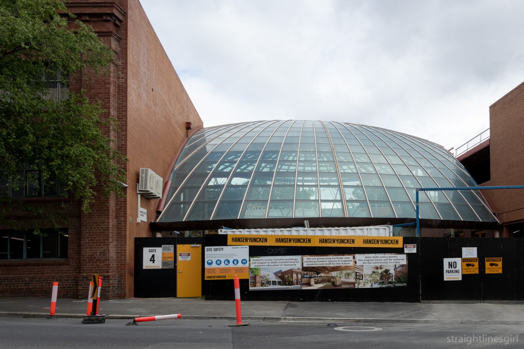 A large glass dome sandwiched betweem two red brick buildings, with construction hoarding along the front and yellow signs bearing the words Hansen Yuncken