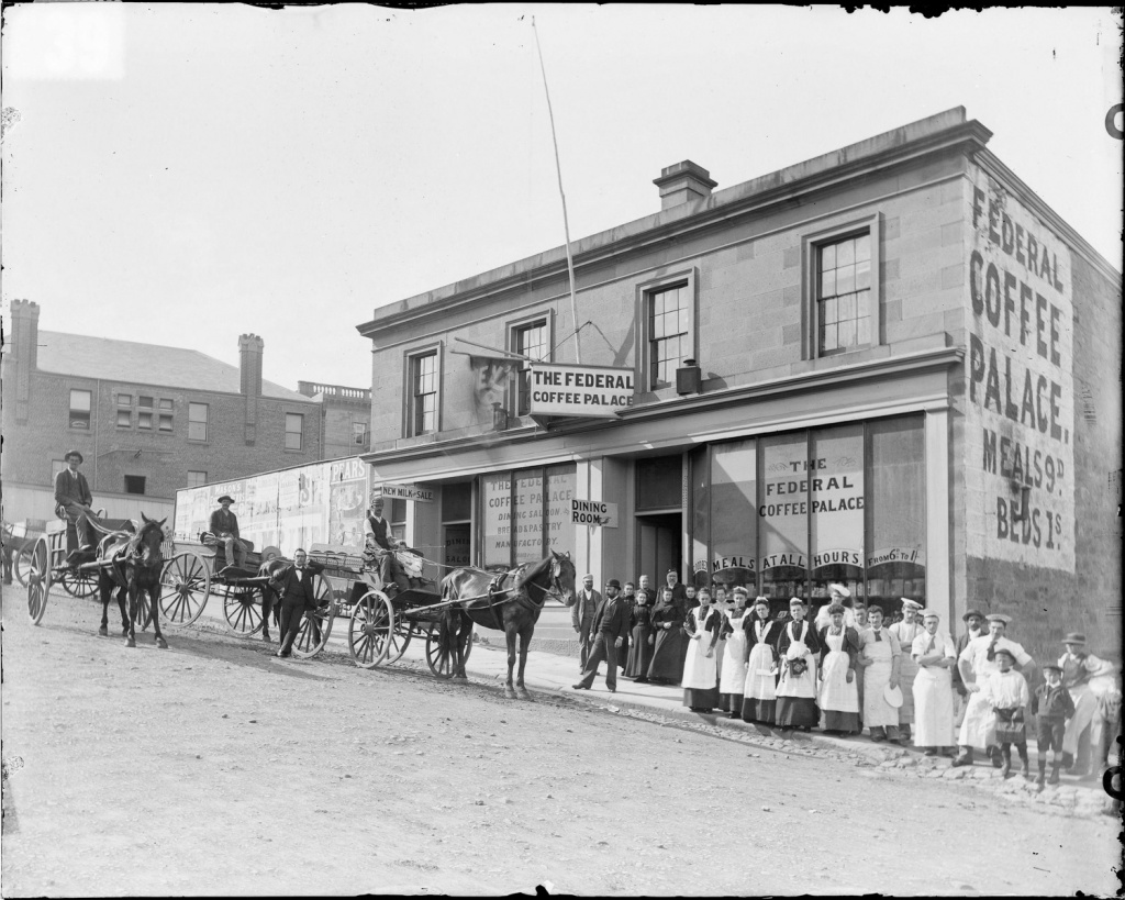 An old black and white photo of the Federal Coffee Palace, a two-storey sandstone building, with a line of people and some horses and carts outside