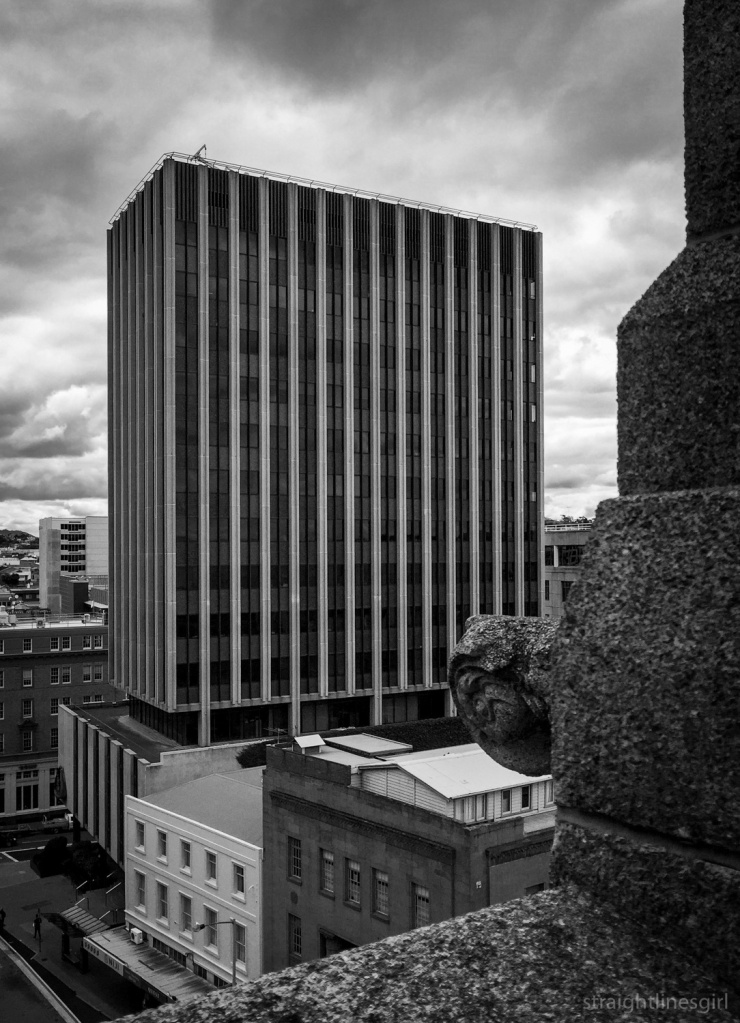 A black and white photo of a large concrete building sitting on a podium, with the edge of another buildin gint he foreground  