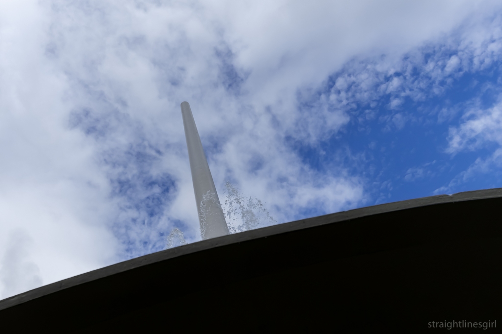 Looking up at a cloudy blue sky from the underside of the fountain. The spire of the fountain soars into the sky