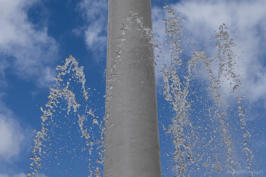 Close up image of water jets against a fountain spire, with blue sky and clouds in the background