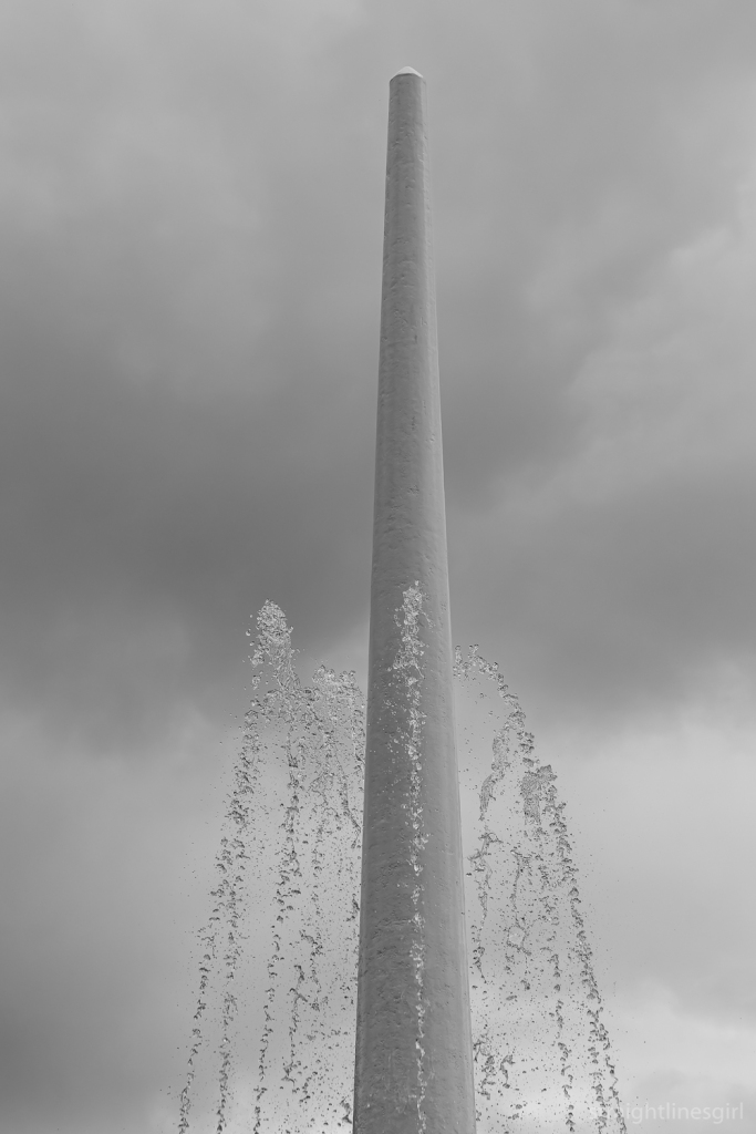 Black and white image of a fountain sire with tall water jets against a cloudy sky