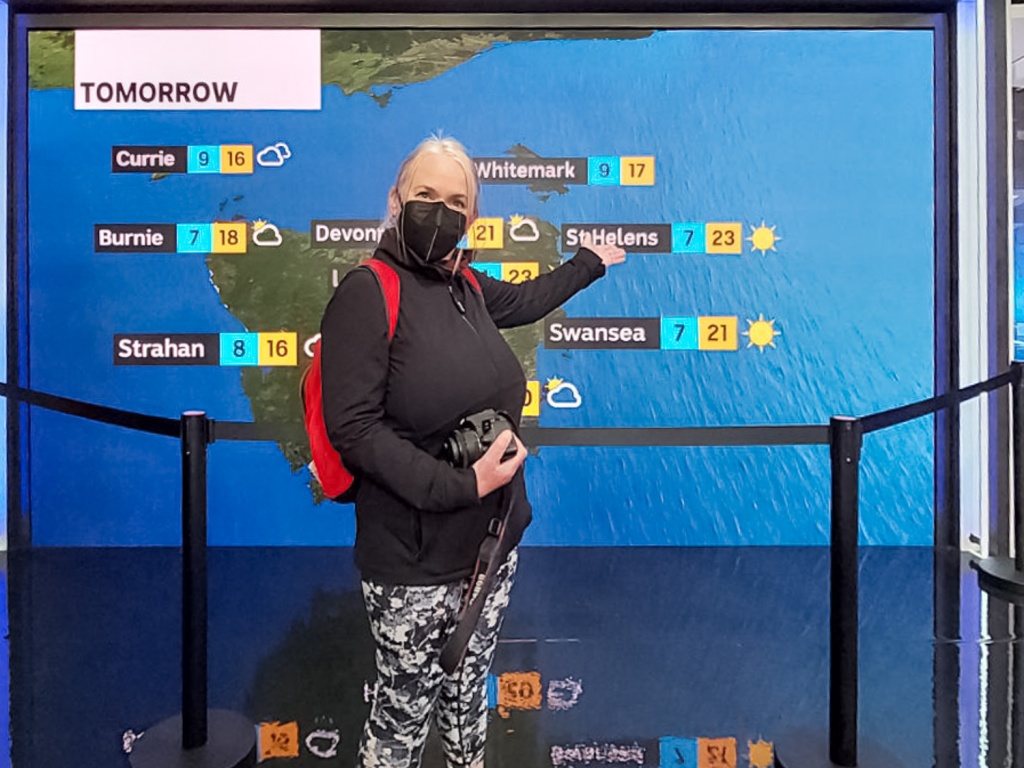 Barb is standing in front oe a map of Tasmania's weather holding out her arm to St Helens. She is wearing floral leggings, a black jacket and a black face mask. She has a camera in her other hand.