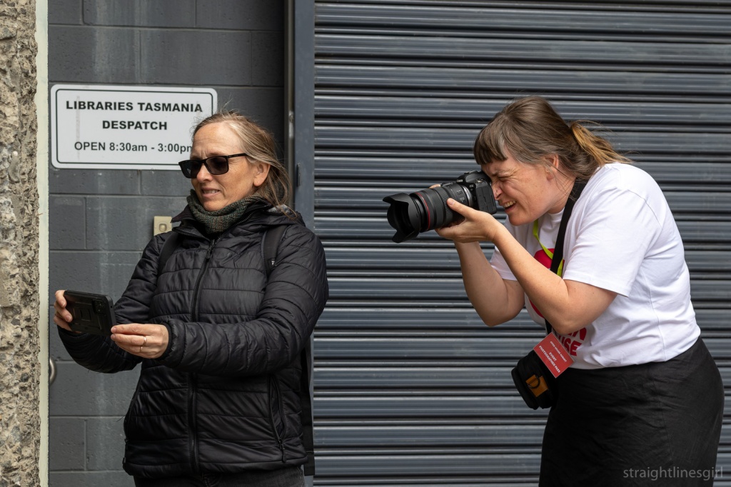 A woman in a white t-shirt is photographing a woman in a black jacket who is photographing the side of a building