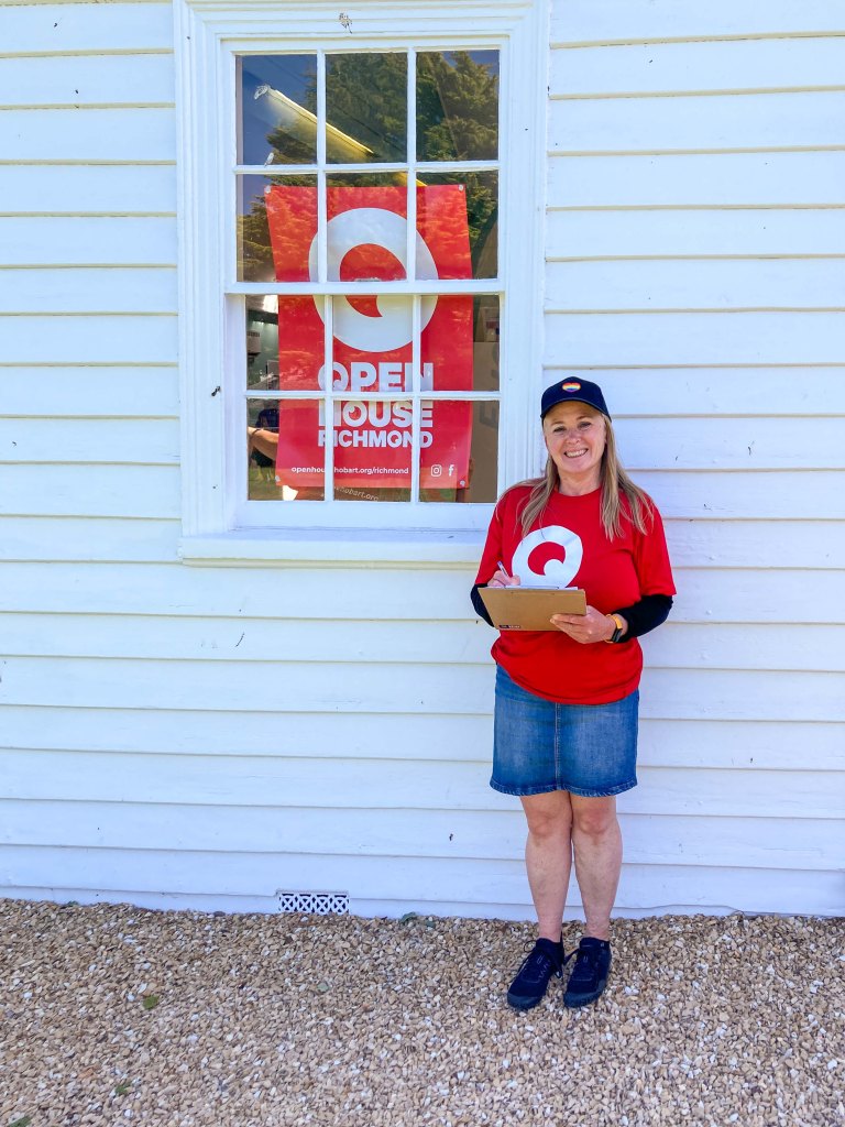Barb is wearing a red Open House t-shirt holding a clipboard standing outside of a white weatherboard building with a red Open House Richmond sign in the window. She is wearing a black cap and a demin mini skirt. 